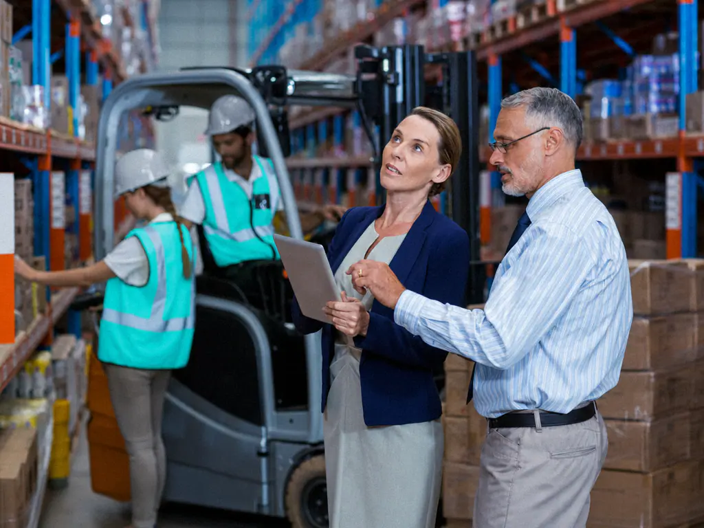 Deux responsables logistiques consultant une tablette dans un entrepôt, devant un chariot élévateur