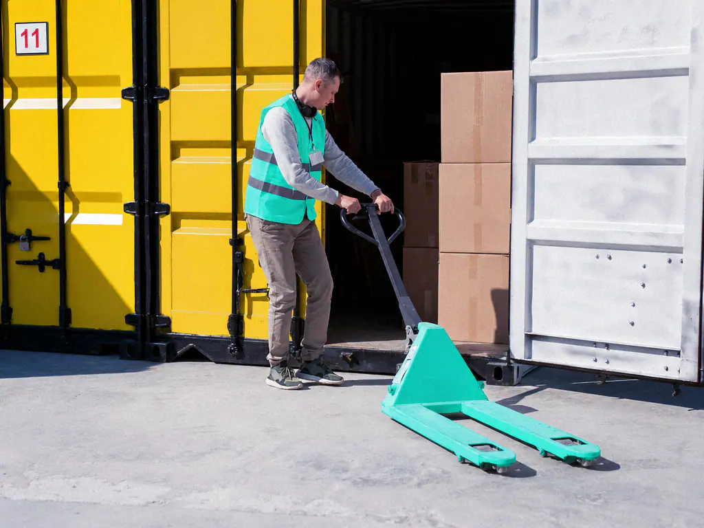 Manutentionnaire en gilet mint chargeant des cartons dans un conteneur jaune avec un transpalette