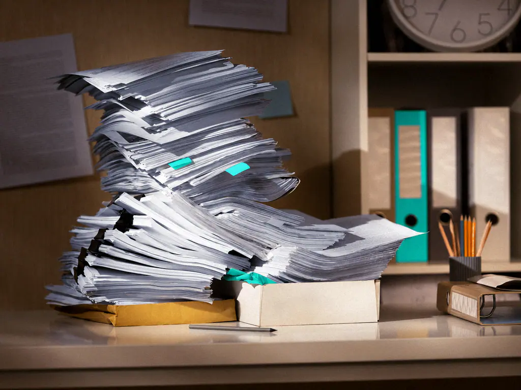 Pile de dossiers papier en attente sur un bureau, à côté des classeurs d’archives