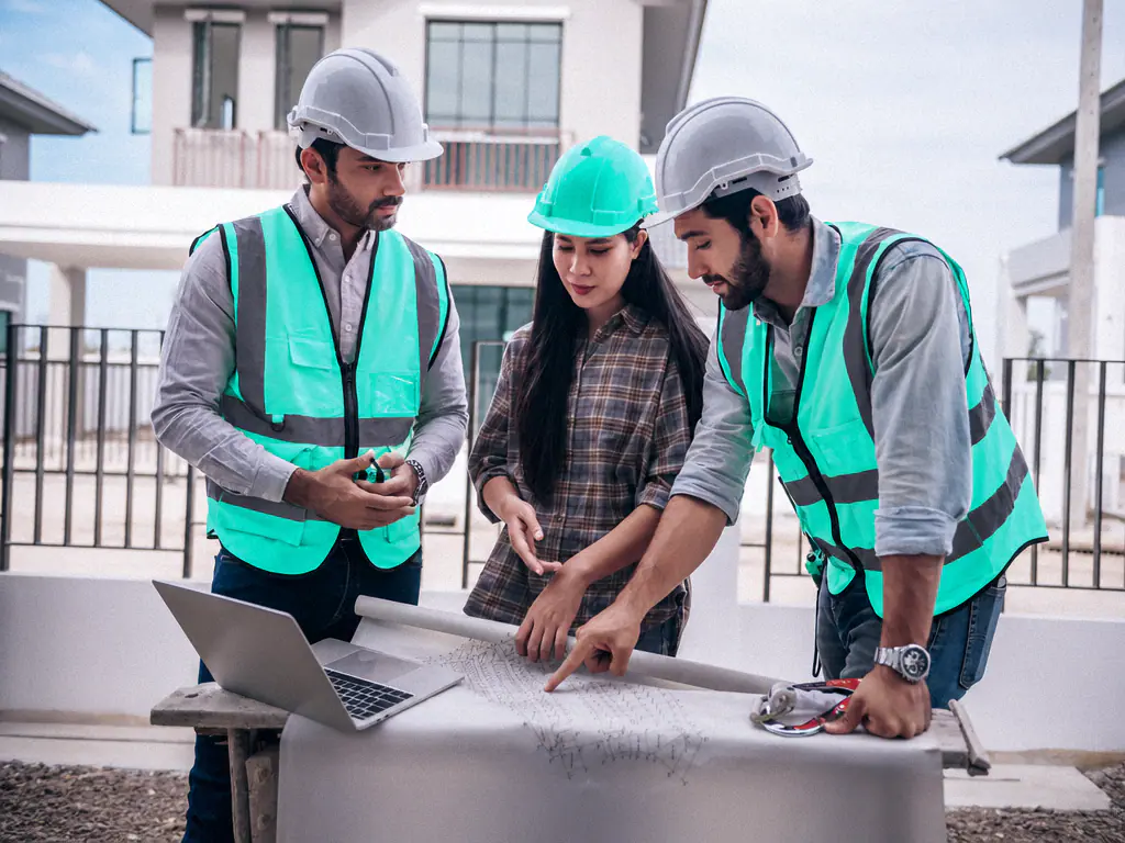 Trois ingénieurs en casque et gilet étudient un plan de construction avec un ordinateur portable sur le chantier