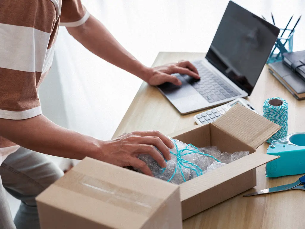 Mains emballant un article protégé dans un carton, ordinateur portable ouvert sur le bureau
