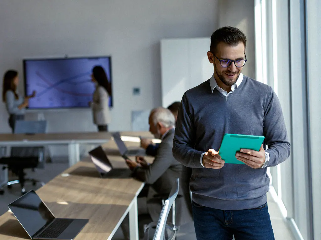 Formateur consultant une tablette dans une salle de formation où des participants travaillent sur ordinateur