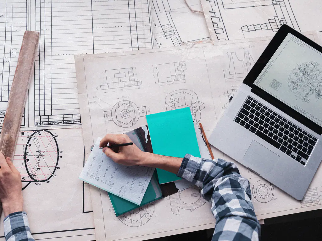 Mains annotant des dessins techniques à côté d’un ordinateur portable ouvert sur un plan