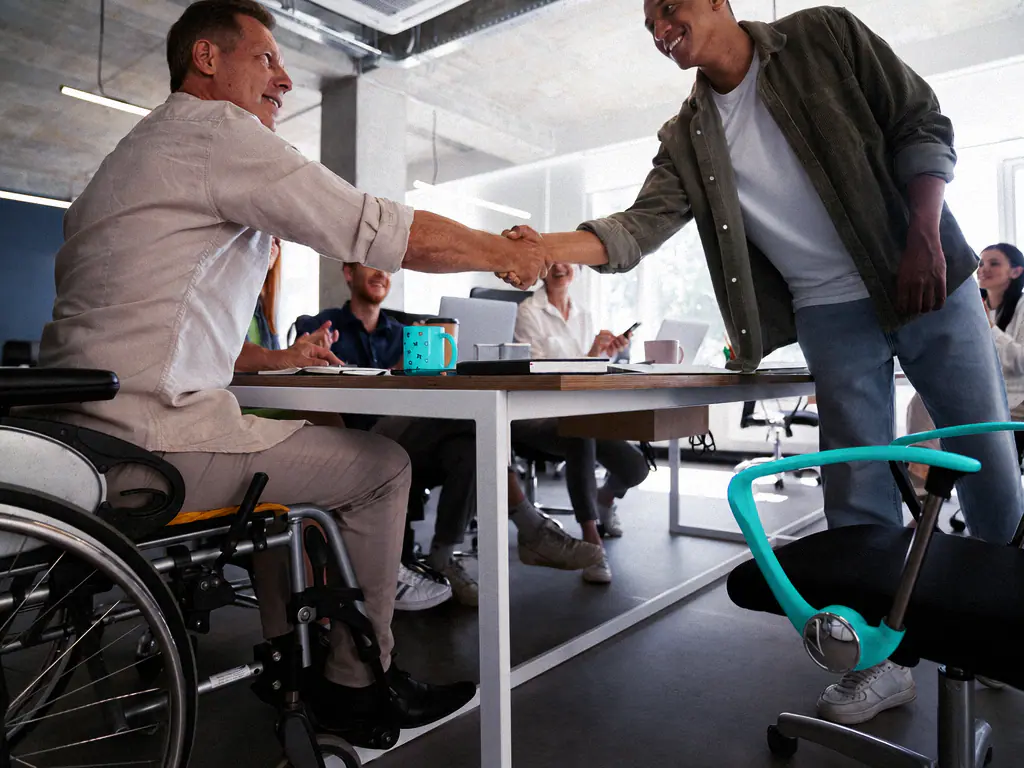 Poignée de main d’accueil entre un nouveau collaborateur et un collègue en fauteuil roulant, autour d’une table de réunion