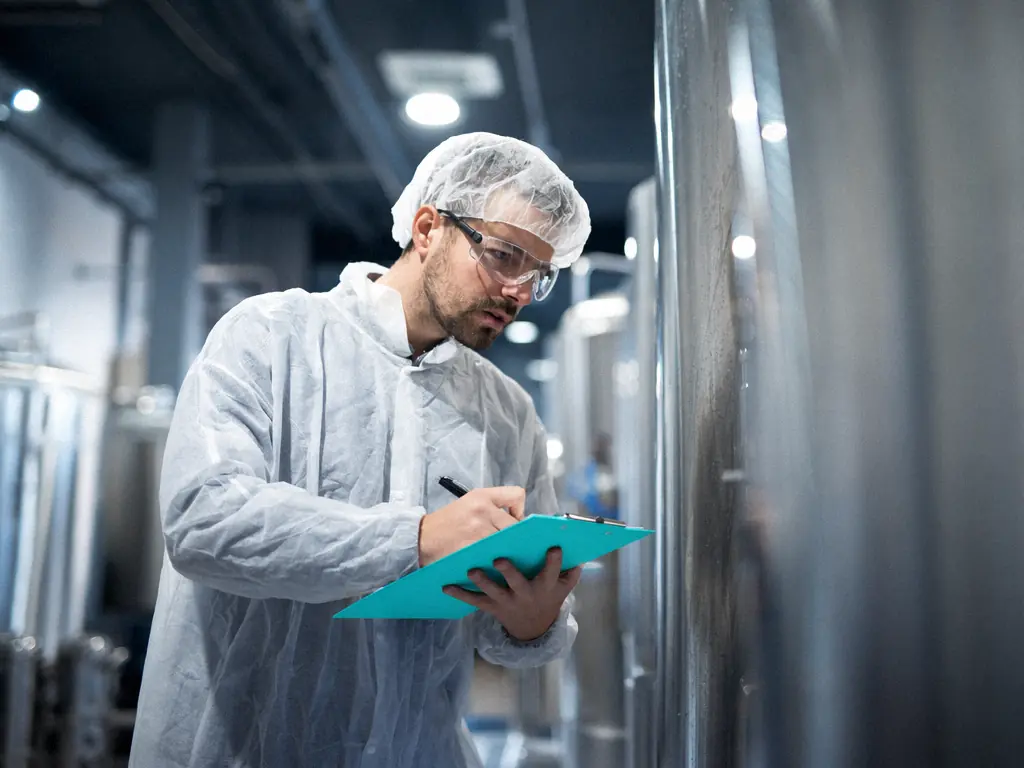 Technicien en tenue de salle propre et lunettes remplissant une fiche de contrôle près d’une cuve inox