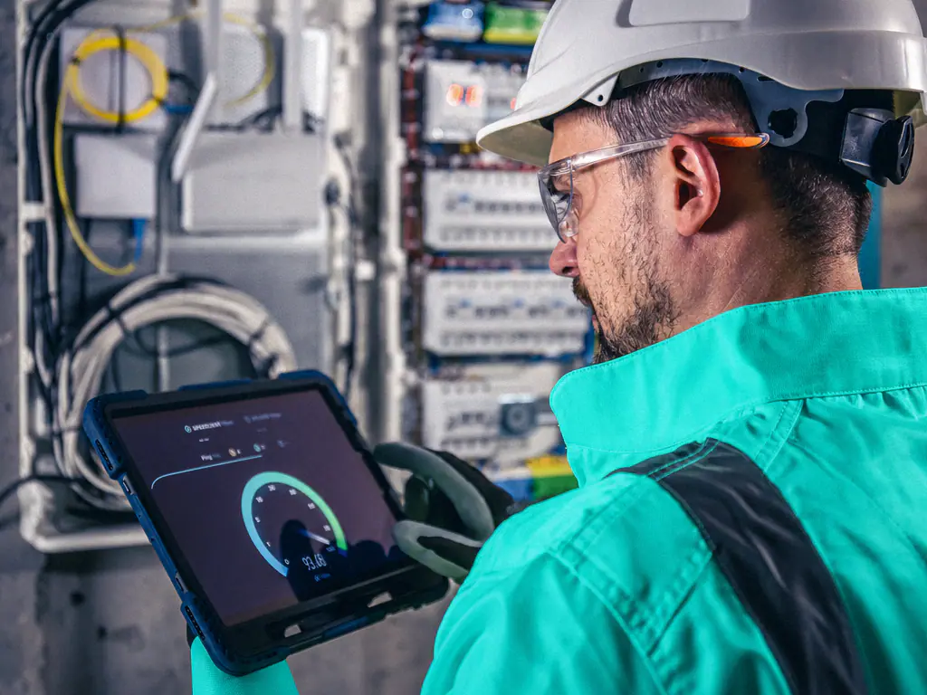Un technicien casqué consulte une tablette devant une baie de télécommunications